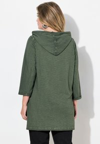 Tunique à capuche verte avec des manches trois quarts, en tissu texturé, et une coupe ample, atteignant le milieu de la cuisse. Design simple avec des poignets retroussés.