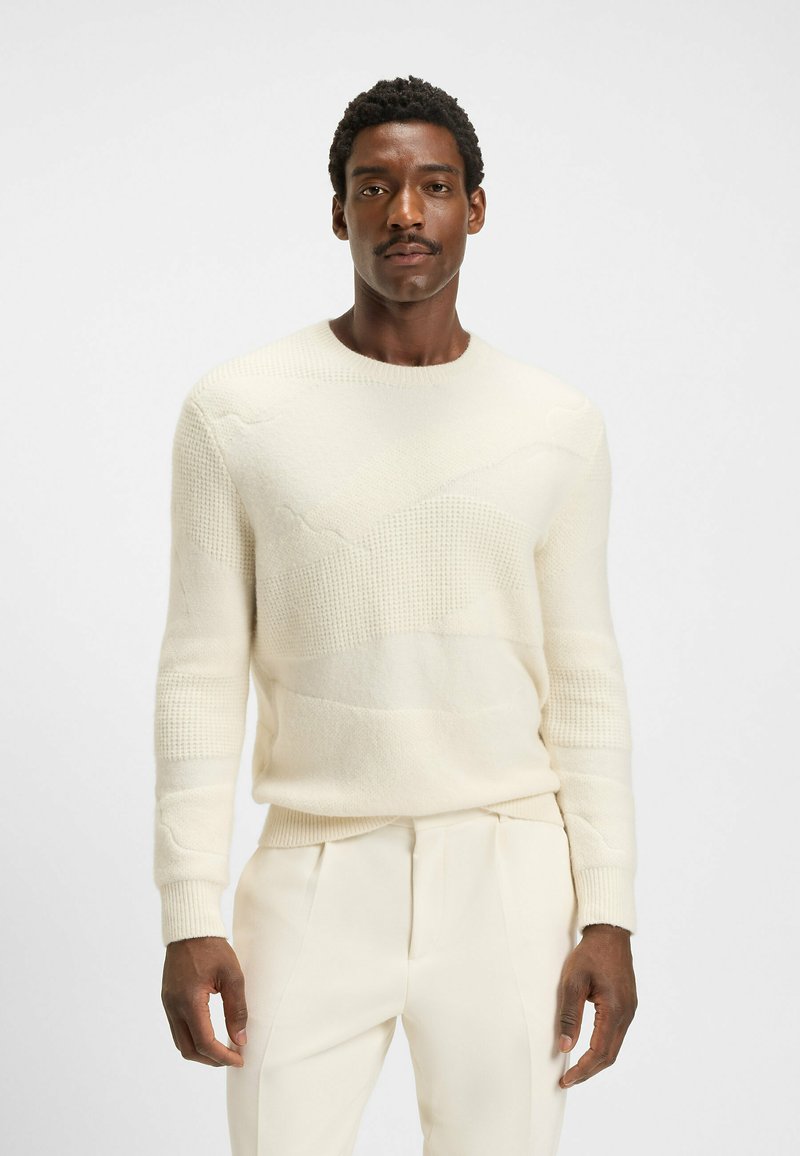 Strikket creme sweater med teksturerede striber og rund halsudskæring, parret med matchende lyse bukser. Blødt, hyggeligt stof med subtile mønstre.