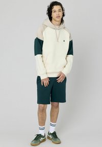 Sweatshirt à capuche crème avec des accents verts et beiges, associé à un short vert foncé. Le modèle porte des chaussettes blanches avec des rayures vertes et des baskets.
