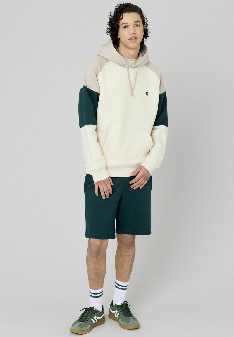 Sweatshirt à capuche crème avec des accents verts et beiges, associé à un short vert foncé. Le modèle porte des chaussettes blanches avec des rayures vertes et des baskets.