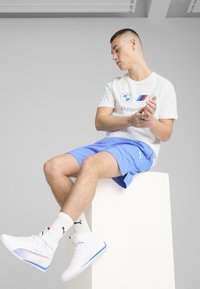 Hvid t-shirt med BMW-logo, blå shorts og hvide højtop sneakers med røde og blå accenter. Model sidder på en lysfarvet sokkel.