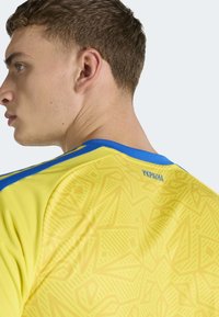 Maglia sportiva gialla con accenti blu, caratterizzata da un motivo geometrico e dalla scritta "УКРАЇНА" sul retro del collo.
