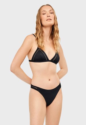 Woman with long wavy blonde hair wearing a black bikini with crochet detailing, standing against a plain light gray background.