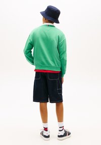 Παιδί που φοράει πράσινο φούτερ, μπλε καπέλο τύπου κάπα, σκούρες τζιν βερμούδες, λευκές κάλτσες και μπλε αθλητικά παπούτσια, στέκεται γυρισμένο με την πλάτη σε λευκό φόντο.