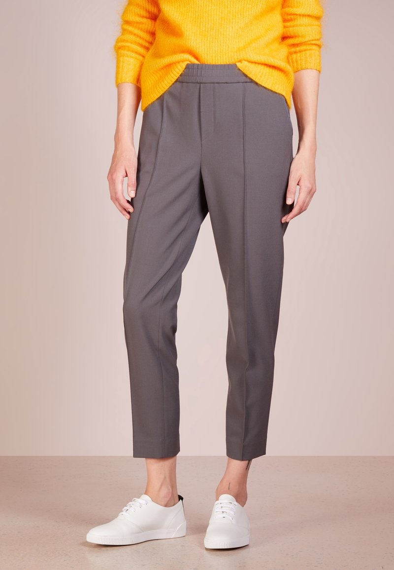 Pantalon gris fuselé avec une texture lisse, taille élastique et plis avant, associé à des baskets blanches sur une surface texturée claire.