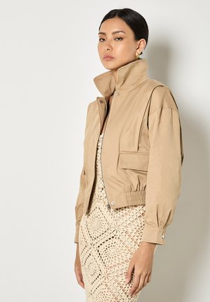 Kobieta w beżowej, krótkiej kurtce z wysokim kołnierzem, nałożonej na kremową sukienkę z wzorem szydełkowym, stojąca na jednolitym jasnym tle.