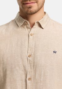 Close-up of a man wearing a beige button-up shirt with a small dark blue embroidered logo on the left chest.