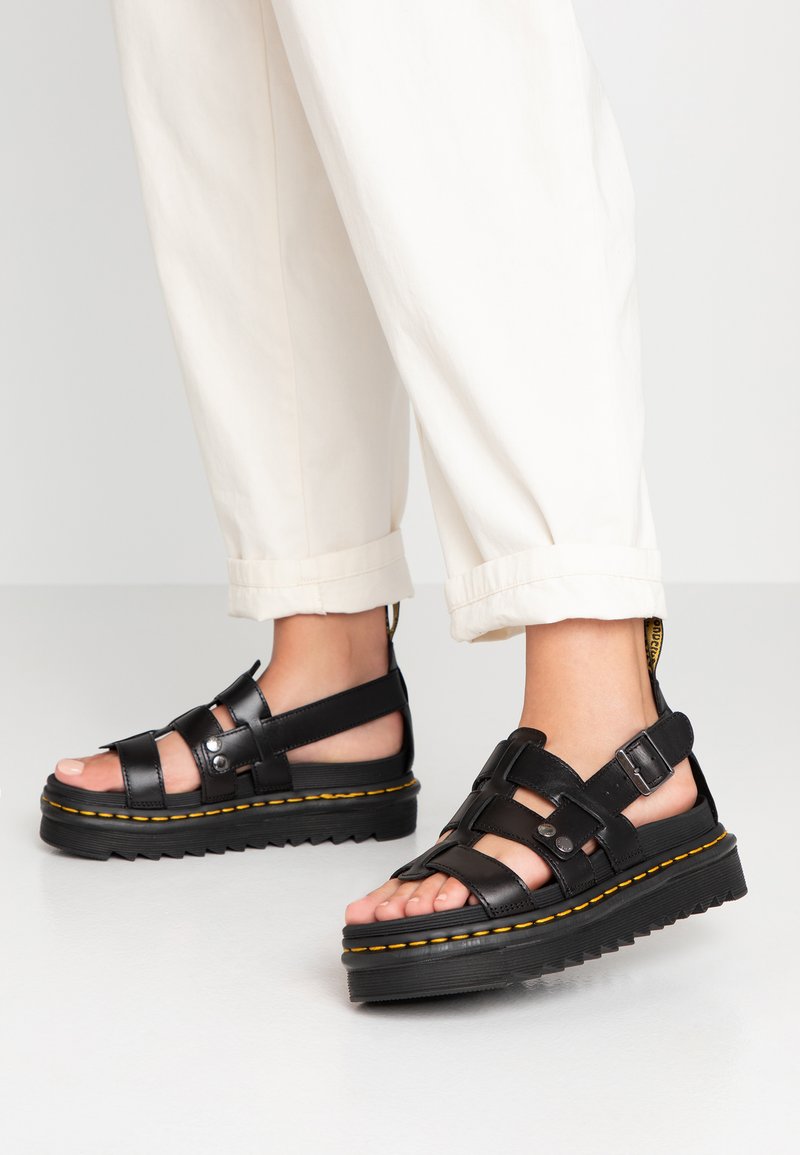 Sandalias de plataforma de cuero negro con correas ajustables, costuras amarillas y una suela de goma texturizada. Llevadas con pantalones crema remangados.