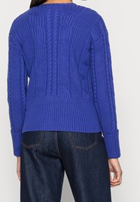 Virkat tröja i livlig blå färg med kabelstickad detalj på baksidan, ribbad nederkant och ärmslut; ihop med mörka denimbyxor.