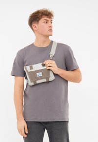 Borsa a tracolla grigia e beige realizzata in tessuto resistente, dotata di tracolla regolabile, tasca frontale e etichetta con marchio.