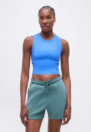 Atletyczna kobieta stojąca, ubrana w niebieski bezrękawnik typu crop top i turkusowe szorty z troczkiem, na jednolitym białym tle.