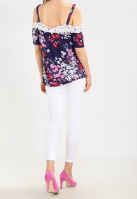Navy floral off-shoulder top with white lace trim, paired with white skinny jeans and pink high-heeled shoes, showcasing vibrant patterns.