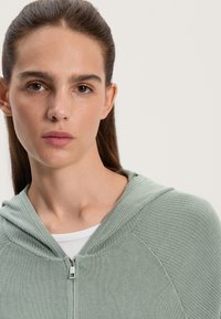 Svetlozelená mikina na zips s rebrovanou textúrou a kapucňou. Nosená cez biely top, má viditeľné švy a malý detail zipsu.