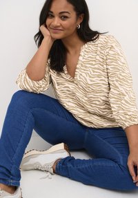 Frau, die auf dem Boden sitzt, mit dem Ellenbogen auf dem Knie, trägt ein beige-weißes Zebra-Muster-Oberteil, blaue Jeans, weiße Sneakers und silberne Creolen.