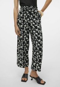 Zwarte wijde broek met een wit bloemenpatroon, hoge taille, een strikceintuur en zijzakken, gecombineerd met zwarte open teen muilen.