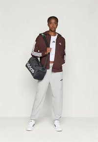 Νέα γυναίκα φορεμένη με ενεργό ένδυμα Adidas: μπορντό φόρμα, λευκό crop top, γκρι αθλητικά παντελόνια, λευκά αθλητικά παπούτσια, κρατώντας μαύρη αθλητική τσάντα Adidas στον ώμο.