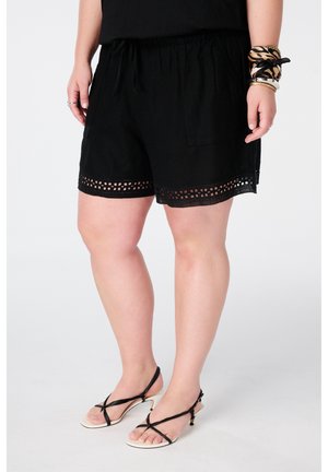 Vrouw draagt zwarte shorts met kanten rand, zwarte mouwloze top, dierenprint en gouden armbanden, en zwart-witte sandalen met bandjes en hakken.
