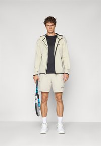 Chaqueta beige clara con detalles en negro, camiseta negra, pantalones cortos beige y zapatillas blancas; sosteniendo una raqueta de tenis azul y negra.