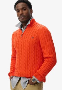 Pomarańczowy sweter z dzianiny w splot warkoczowy, z wysokim kołnierzem i zamkiem, noszony na niebieskiej i białej bluzce w paski z guzikami. Złoty zegarek zdobi nadgarstek.