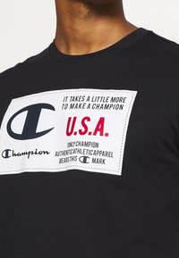 Černé tričko s bílou obdélníkovou nášivkou s textem značky "Champion", velkým logem "C", nápisem "U.S.A." v červené barvě a motivačním sloganem.