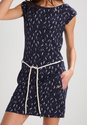Femme portant une robe bleu marine à manches courtes avec un imprimé hippocampe blanc et une ceinture en corde blanche, une main dans la poche, sur fond uni.