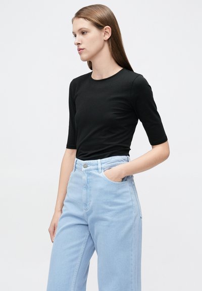 Γυναίκα με μακριά καστανά μαλλιά φοράει μαύρο τοπ με μανίκια μέχρι τον αγκώνα και ανοιχτόχρωμο τζιν με ψηλή μέση, στέκεται με το δεξί της χέρι στην τσέπη.