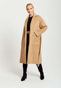 Long, tan coat made of wool blend fabric, featuring a relaxed fit, notched lapel, and two front pockets. Paired with black pants and heels.