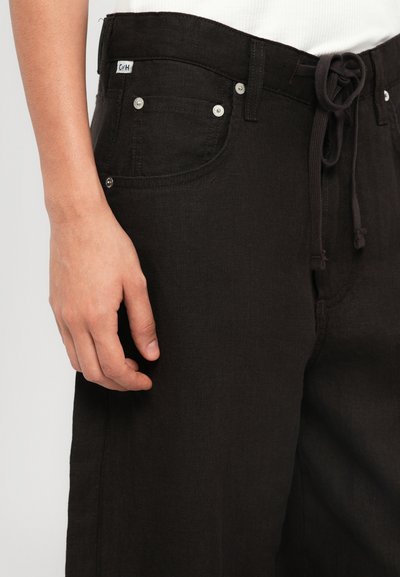 Pantalones de mezclilla negros con cintura ajustable con cordón, bolsillos frontales y detalles en hardware plateado, mostrados sobre un fondo claro.