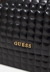 Sac à main noir matelassé avec une finition brillante, de forme rectangulaire et arborant un logo "GUESS" doré à l'avant.