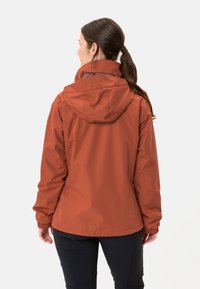 Vrouw met donker haar in een paardenstaart draagt een roestkleurige hooded jas en zwarte broeken, staand met de rug naar de camera tegen een effen achtergrond.