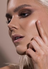 Primo piano di un volto con pelle leggermente abbronzata, un lieve illuminante, ombretto scuro e un anello al setto nasale dorato. Una mano sfiora delicatamente la guancia.