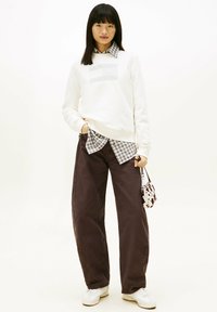 White sweatshirt with gray logo, layered over a plaid button-up shirt. Dark brown wide-leg pants and white sneakers. Small patterned handbag.