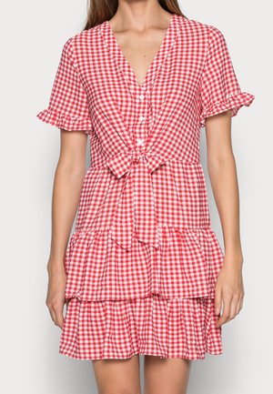 Kobieta ubrana w czerwoną i białą sukienkę w kratę z krótkim rękawem, zapinaną na guziki, z wiązaniem z przodu i warstwową falbanianą spódnicą.