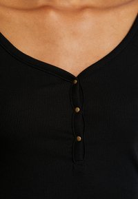 Musta ribbineuloksinen toppi, jossa V-aukko ja kolme messinkinväristä nappia edessä. Pinta on teksturoitu ja malli istuva.