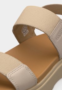 Μπεζ σανδάλι με λουριά από υφασμάτινη υφή και επενδεδυμένη σόλα, με κλείσιμο βέλκρο για ρυθμιζόμενη εφαρμογή.