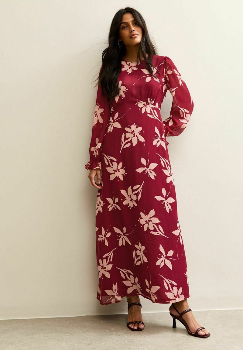 Vrouw met lang zwart haar die een lange, rode maxi-jurk met grote lichte bloemenprints en zwarte hoge hakken draagt.
