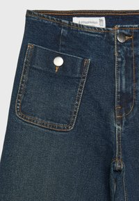 Jean en denim bleu foncé avec des coutures orange, une braguette à boutons sur le devant, et une poche carrée à bouton métallique sur le côté gauche, montrant une étiquette de taille à l’intérieur.