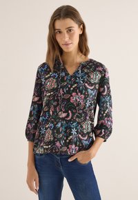 Jeune femme portant une blouse noire à fleurs avec des manches trois-quarts et un jean bleu, debout avec une main dans la poche, sur un fond uni.