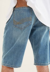 Pantalones cortos de mezclilla en azul claro con un corte holgado, que presentan un bolsillo trasero visible con costuras decorativas en forma de ola en dorado.