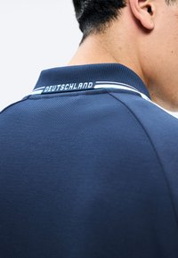 Man draagt een marineblauw sportshirt met "DEUTSCHLAND" gedrukt op de achterkant van de kraag, toont de rechterkant van zijn hoofd en nek.
