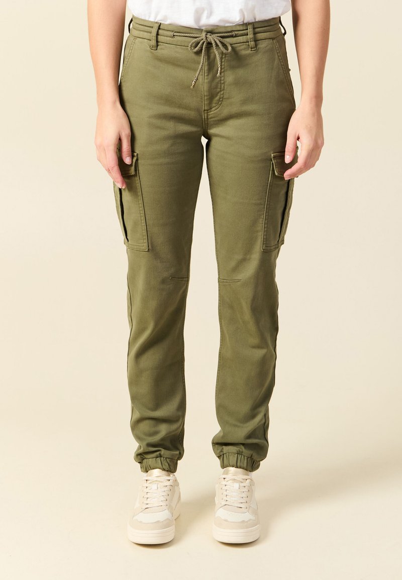 Femme portant un pantalon cargo vert olive avec taille à cordon de serrage et poignets élastiques, assorti à des baskets blanches et une chemise blanche rentrée.