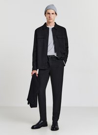 Schwarzes Hemd zum Knöpfen über einem grauen T-Shirt, kombiniert mit schwarzen strukturierten Hosen und schwarzen Stiefeln. Eine graue Strickmütze wird getragen. Hält einen schwarzen Pullover.