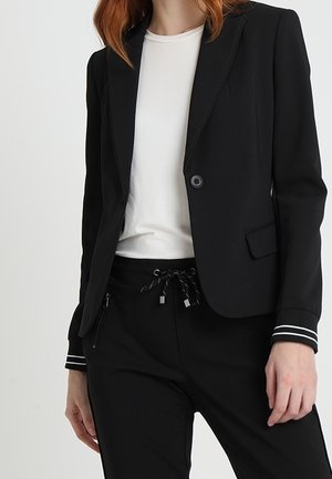 Mujer vistiendo un blazer negro ajustado con puños a rayas blancas, una camiseta blanca y pantalones negros con cordón y bolsillos con cremallera.