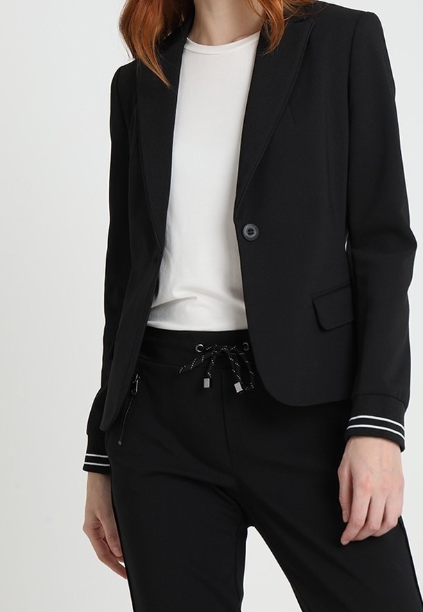 Femme portant un blazer noir ajusté avec des poignets à rayures blanches, un t-shirt blanc et un pantalon noir avec cordon de serrage et poches zippées.