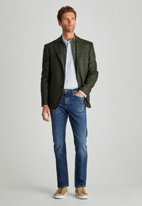 Blazer de tweed verde oscuro sobre una camisa de rayas azul claro, combinado con jeans azules y zapatillas beige, con un corte ajustado y dos bolsillos.