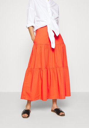 Nainen, jolla on kirkkaan oranssi kerroksittainen maksihame, valkoinen napillinen paita solmittuna vyötäröltä ja mustat liukuva sandaalit, joissa on solkikoriste.