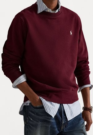 Mann trägt einen kastanienbraunen Pullover über einem blau gestreiften Hemd, Jeans, linke Hand in der Tasche, Uhr am rechten Handgelenk.