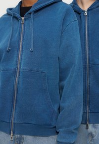 Δύο άτομα φορούν μπλε φούτερ με φερμουάρ και μπροστινές τσέπες, καθώς και ανοιχτόχρωμα μπλε τζιν, εμφανίζονται από τον λαιμό μέχρι τους γοφούς.