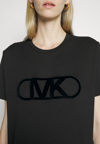 Camiseta de algodón negra de manga corta, con un logo en relieve de terciopelo negro "MK" en el frente. Cuello redondo sencillo, ajuste relajado.