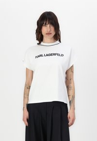 Balta kokvilnas T-kreklā ar īsām piedurknēm, apaļu kakla izgriezumu un drukātu logotipu "KARL LAGERFELD" treknā melnā tekstā uz priekšpuses.